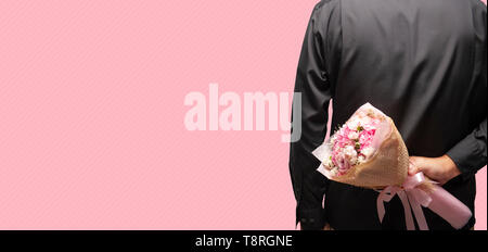 Lo sposo che indossa la maglia nera tenendo bouquet dietro la schiena su sfondo rosa, uomo nascondendo fiori a sorpresa, anniversario buon compleanno. Foto Stock