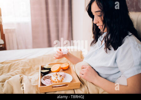 Bella awesome dai capelli scuri donna avente una gustosa prima colazione a letto con il suo accogliente camera da letto. Vista laterale foto della ragazza in blu pigiami a strisce di mangiare sw Foto Stock