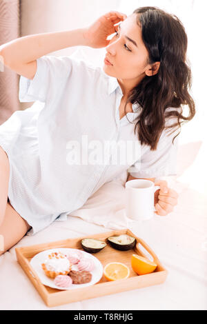 Close up ritratto di bella donna giovane in maglietta blu guardando alla finestra e godersi la giornata di sole e di bere il caffè. Rilassati ragazza sognante in posa di letto, Foto Stock