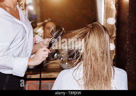 Close up professionale di parrucchiere a spazzola e asciugacapelli styling lunghi capelli biondi del suo client femmina. Una sana lucida i capelli della donna dopo capelli tr Foto Stock