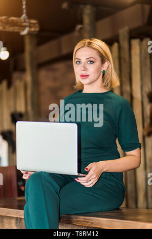 Allegro elegante bionda business donna, amministratore indossando color smeraldo suit seduta sul banco della reception in legno azienda laptop, divertendosi in workpl Foto Stock