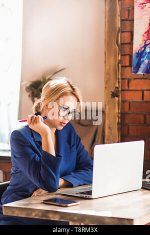 Giovane attraente femmina caucasica blogger digitando nuovi post durante l udienza presso il cafe con computer portatili e smart phone sul tavolo. Bella bionda business w Foto Stock