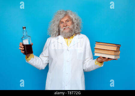 Vecchio con i capelli lunghi uomo che indossa Doctor's vestaglia studio isolato sulla parete blu in piedi tenendo la bottiglia di whisky in una mano e la pila di libri in un altro gabinetto Foto Stock