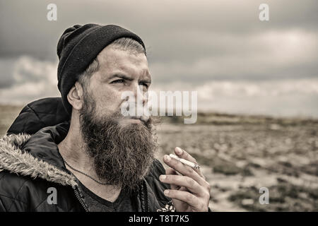 Barbuto hipster con barba lunga il fumo di sigarette in hat su sfondo naturale, cattiva abitudine e la tossicodipendenza, lo stile di vita Foto Stock