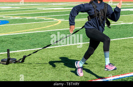 Una scuola di velocista femmina è in esecuzione mentre si tira una slitta con un peso su di esso per la resistenza alla formazione, su un tappeto erboso verde campo. Foto Stock