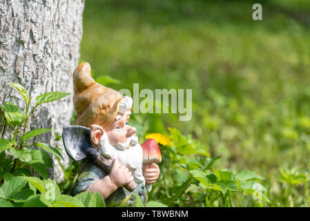 La figura di uno gnomo da giardino con una pala in corrispondenza di un albero in erba verde. Close-up. Copia dello spazio. Foto Stock