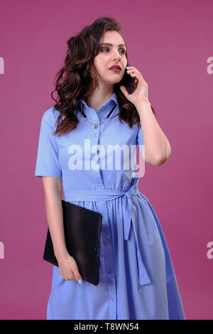 Foto di business donna in vestito blu holding appunti con file e chiamare con il cellulare su sfondo rosa Foto Stock