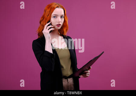 Foto di donna di successo 20s holding appunti con i file di Office e parlando al telefono cellulare su sfondo rosa Foto Stock