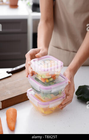 Donna che mantiene pila di contenitori in plastica con verdure fresche per congelamento a tavola in cucina Foto Stock