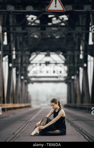 La ballerina è seduta e la sua legatura pointe scarpe tra le rotaie sulla strada contro lo sfondo di supporti metallici. Foto Stock