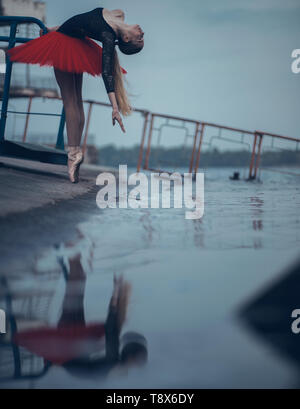Ballerina ballare sulla costa del fiume in un nero e rosso tutu e il suo riflesso nell'acqua. Foto Stock