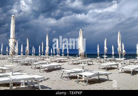 Buia e tempestosa sky, con nuvole grigie sul mare blu e un po' di spiaggia con sedie a sdraio e chiuso ombrelloni bianchi in primo piano Foto Stock