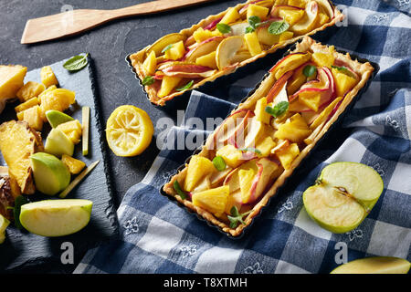 Deliziosa ananas e apple sweet tart rettangolare Tart padelle su un tavolo in cemento con carta asciugatutto e ingredienti, vista orizzontale dal di sopra, Foto Stock