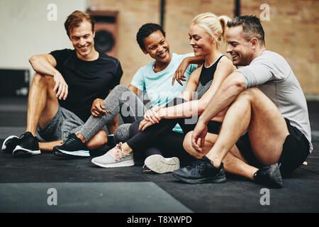 Gruppo di amici sorridente in sportswear parlare insieme seduti su un pavimento palestra dopo gli allenamenti Foto Stock