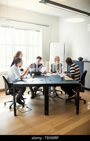 Diversi gruppi di giovani designer a discutere di lavorare insieme seduti attorno a un tavolo per riunioni in un ufficio Foto Stock