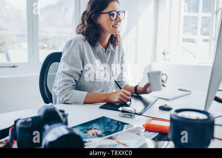 Giovane donna seduta alla scrivania, la modifica delle immagini sul computer. Fotografo femmina il ritocco di foto in ufficio utilizzando tavoletta grafica e penna digitale. Foto Stock