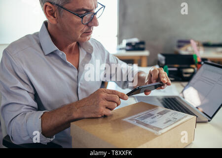 Imprenditore maturo seduto alla sua scrivania e la scansione di una scatola di consegna con il suo telefono cellulare. L'uomo della vendita di prodotti online, la scansione del codice a barre con il suo m Foto Stock