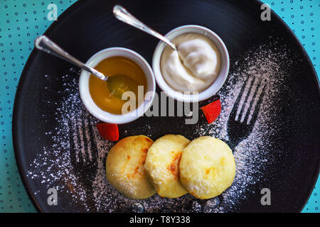 Una sana colazione cottage cheesecake sirniki russo con miele e panna acida per la colazione su sfondo blu. Foto Stock