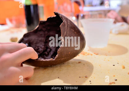 Un uovo di pasqua è stato aperto, rotto di guscio di cioccolato è sul tavolo di appoggio nel suo involucro. Un uomo caucasico la mano taglia un pezzo di cibo. 30 Foto Stock