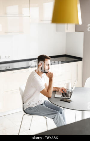 Felice uomo bello freelancer seduto al tavolo della cucina, lavorando sul computer portatile Foto Stock
