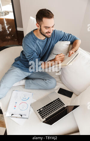 Attraente giovane uomo relax su un lettino a casa, lavorando con computer portatile e documenti Foto Stock
