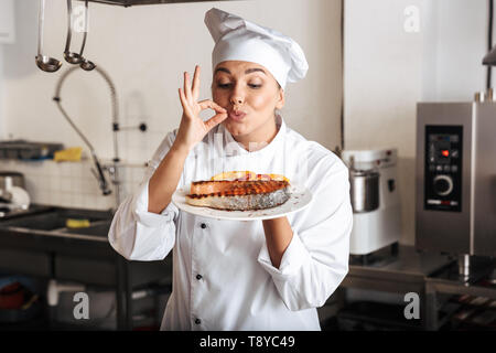 Immagine della bella donna chef indossando bianco uniforme tenendo la piastra con grigliate di pesce in cucina presso il ristorante Foto Stock