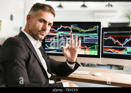 Immagine dell'imprenditore unshaved 30s indossare tuta di lavoro di Office sul computer con grafica e grafici a schermo Foto Stock