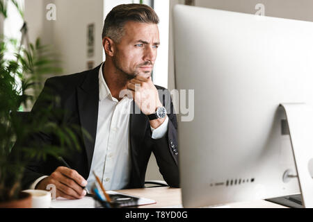 Immagine dell'imprenditore fiducioso 30s indossare tuta lavoro in ufficio e cerca sul computer Foto Stock