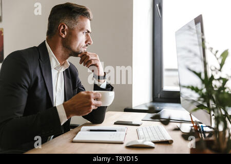 Immagine dell'imprenditore europeo 30s indossare tuta di bere il caffè mentre si lavora sul computer in ufficio Foto Stock
