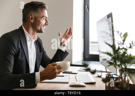 Immagine di gioiosa imprenditore 30s indossare tuta di bere il caffè mentre si lavora sul computer in ufficio Foto Stock