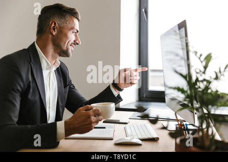 Immagine della bella imprenditore 30s indossare tuta di bere il caffè mentre si lavora sul computer in ufficio Foto Stock