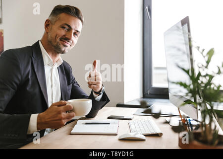 Immagine di gioiosa imprenditore 30s indossare tuta di bere il caffè mentre si lavora sul computer in ufficio Foto Stock
