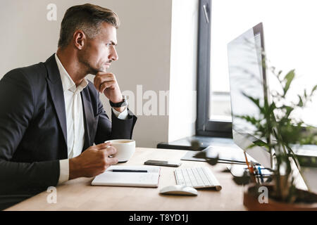 Immagine dell'imprenditore concentrato 30s indossare tuta di bere il caffè mentre si lavora sul computer in ufficio Foto Stock