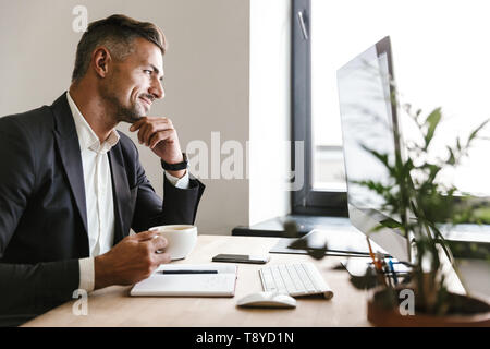 Immagine dell'imprenditore unshaved 30s indossare tuta di bere il caffè mentre si lavora sul computer in ufficio Foto Stock
