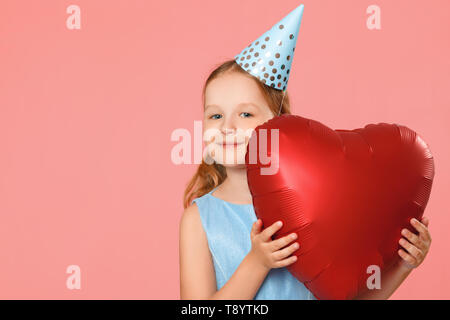 Felice bambina in una capsula contiene un grande cuore palloncino a forma di. Ritratto di un bambino su un sfondo rosa. Copia dello spazio. Foto Stock