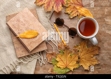 Composizione con tazza di tè aromatici, caldi plaid, libri e foglie di autunno su sfondo di legno Foto Stock