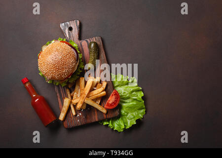 Hamburger fatti in casa con ingredienti di manzo, pomodori, lattuga, formaggio, cipolla, i cetrioli e le patatine fritte sul tagliere e rusty sfondo. Vista dall'alto. Foto Stock