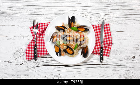 Riso fritto con frutti di mare cozze, gamberi e basilico in una piastra con asciugamano, coltello e forchetta su bianco incrinato un tavolo di legno. Vista dall'alto. Foto Stock