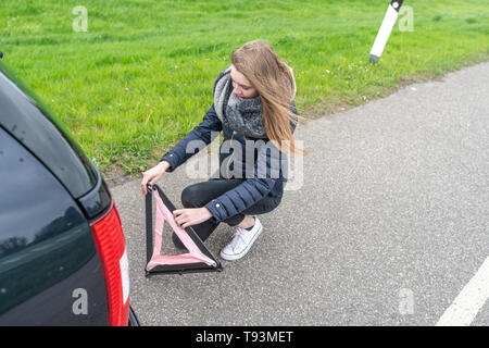 Una giovane donna si inginocchia dietro la vettura e monta il triangolo di avvertenza Foto Stock
