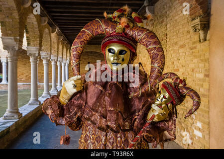 Ritratto di un masculin persona mascherato in una bella creative costume di Arlecchino, ponendo in un cortile con archi e pilastri, celebrando il veneziano Foto Stock