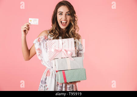 Immagine di donna allegra 20s nel vestire tenendo la carta di credito e scatole con acquisto, isolate su sfondo rosa Foto Stock