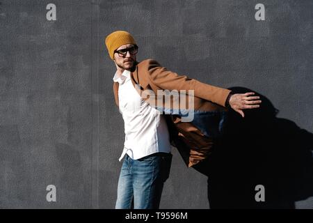 Ritratto di bello uomo barbuto vestito in abiti alla moda in piedi sulla strada contro il muro grigio sfondo Foto Stock