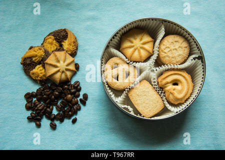 I biscotti con lo zucchero in una scatola rotonda e tre giacenti su una luce blu tovaglia con i chicchi di caffè Foto Stock