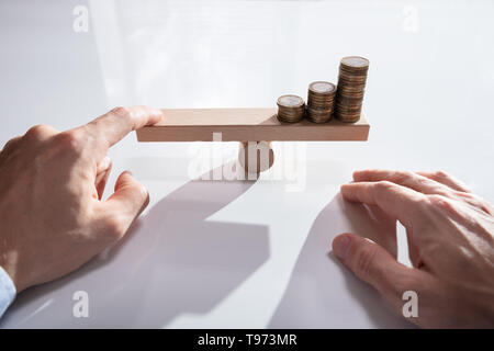 Close-up di un imprenditore la mano di bilanciamento delle monete impilate su altalena in legno con il dito sulla scrivania Foto Stock