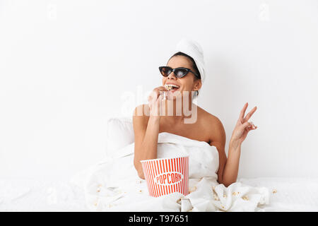 Allegro giovane donna seduta a letto dopo la doccia avvolto in una coperta, indossando occhiali da sole, mangiare popcorn Foto Stock