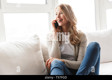 Giovane e bella donna bionda relax su un lettino a casa parlando al telefono cellulare Foto Stock
