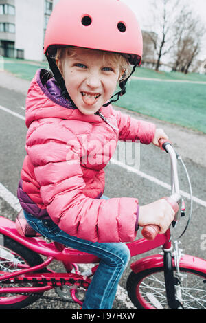 Sorridente preschooler caucasica ragazza rosa di equitazione Bicicletta bici nel casco su strada nel cortile esterno sulla giornata di primavera. Bambino stagionale nozione di attività. Un sano Foto Stock