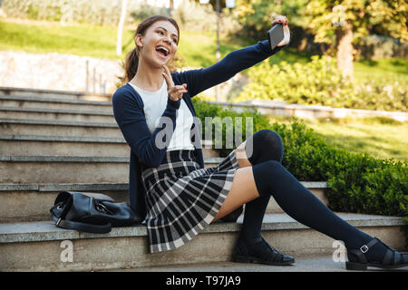 Allegro scuola giovane ragazza seduta all'aperto su scala, utilizzando il telefono cellulare, tenendo selfie Foto Stock