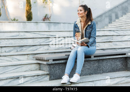 Sorridente giovane donna in giacca seduta su una panchina all'aperto, la lettura di libro, tenere tazza di caffè da asporto Foto Stock