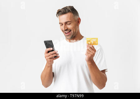 Immagine dell uomo unshaved 30s indossando t-shirt casual tenendo lo smartphone in plastica e carta di credito isolate su sfondo bianco Foto Stock